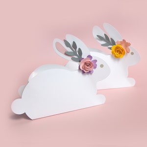 May include: Two white paper bunny decorations with floral accents. One bunny has a yellow flower, the other has a pink flower. The bunnies are set against a soft pink background, perfect for Easter or spring decor.