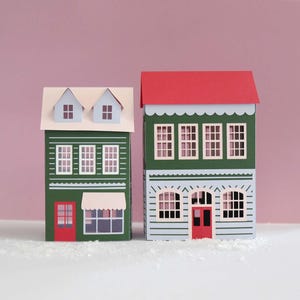 May include: Two paper houses, one green with a white roof and blue dormers, the other green with a red roof. Both have white window frames and red doors. The houses are on a white surface against a pink background. The houses are decorative.