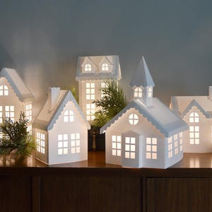Op de afbeelding: Een verzameling van vijf witte papieren huisjes als lantaarns met uitgesneden ramen en deuren, van binnenuit verlicht voor een zachte gloed. De bouwwerken hebben diverse dakstijlen, waaronder puntgevels en een torenspits, met dennentakken.