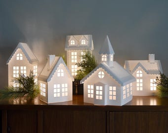 May include: A collection of five white paper house luminaries with cut-out windows and doors, illuminated from within to emit a soft glow. The structures feature various roof styles, including gables and a steeple, accented by green pine sprigs.