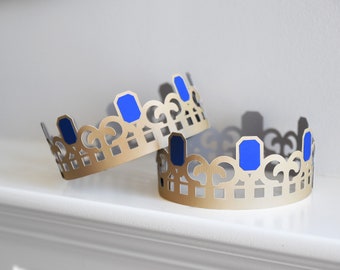 May include: Two gold crowns with blue gem accents. The crowns are made of paper and have a delicate, intricate design.