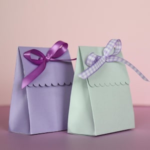 May include: Two small gift bags, one lavender and one mint green, are displayed against a pink background. Each bag is adorned with a decorative ribbon bow. The lavender bag has a solid purple ribbon, while the mint green bag features a checkered ribbon.