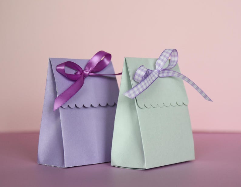 May include: Two small gift bags, one lavender and one mint green, are displayed against a pink background. Each bag is adorned with a decorative ribbon bow. The lavender bag has a solid purple ribbon, while the mint green bag features a checkered ribbon.