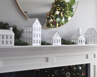 May include: A collection of white paper houses of varying sizes, arranged on a white mantelpiece. The houses have multiple windows and are decorated with greenery. A Christmas tree is reflected in a gold-framed mirror in the background.