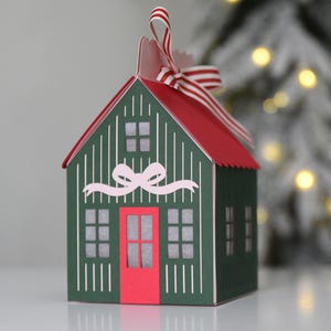 Puede incluir: Una caja de regalo con forma de casa en verde oscuro con techo rojo y puerta roja. El exterior presenta rayas verticales blancas, un gráfico de lazo rosa claro y ventanas recortadas con fondo translúcido. Una cinta a rayas rojas y blancas está atada en la parte superior.