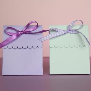 May include: Two small, square gift boxes. One is lavender with a solid purple ribbon, the other is mint green with a gingham ribbon. Both boxes have a scalloped edge along the top.