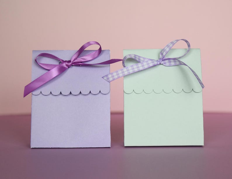 May include: Two small, square gift boxes. One is lavender with a solid purple ribbon, the other is mint green with a gingham ribbon. Both boxes have a scalloped edge along the top.