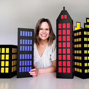 Puede incluir: Cuatro recortes de edificios de cartón negro con ventanas de colores. Los edificios tienen ventanas amarillas, rojas y azules. Una persona sonríe detrás de los edificios.
