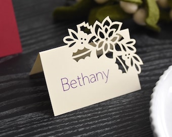 May include: Ivory place card with a floral and holly leaf cutout design. The name "Bethany" is printed in purple. The card is folded and stands upright on a dark wood surface, with a red card and white plate in the background.