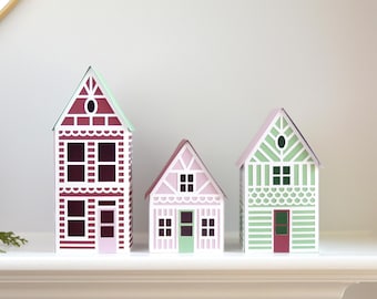 Op de afbeelding: Drie papieren huizen in verschillende kleuren en ontwerpen. Het grootste huis is rood met witte versieringen en een groen dak. Het middelste huis is roze met witte versieringen en een groen dak. Het kleinste huis is groen met witte versieringen en een rode deur.