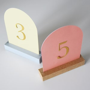 Op de afbeelding: Twee tafelnummerhouders, een gele en een roze, met gouden nummers 3 en 5. De houders zijn gemaakt van karton en hebben een eenvoudig ontwerp.