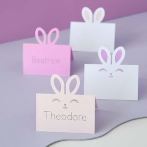 May include: Four pastel-colored place cards with bunny ear cutouts. Two are pink, one is light pink, and one is white. The pink cards have names printed on them. The white cards have a bunny face design. The background is a gradient of purple.