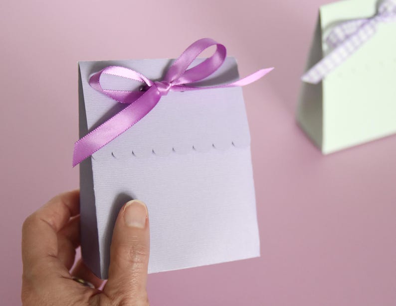 May include: A light purple gift box with a scalloped edge and a purple satin ribbon tied in a bow. The box is held by a hand against a pink background. Another light green box with a white and purple ribbon is in the background.