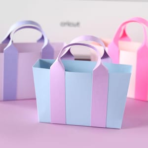 Könnte beinhalten: Drei pastellfarbene Papiertragetaschen. Die Taschen sind in den Farbtönen Lavendel, Hellblau und Rosa gehalten und haben passende vertikale Streifen und Griffe. Die Taschen sind auf einer hellvioletten Oberfläche angeordnet.