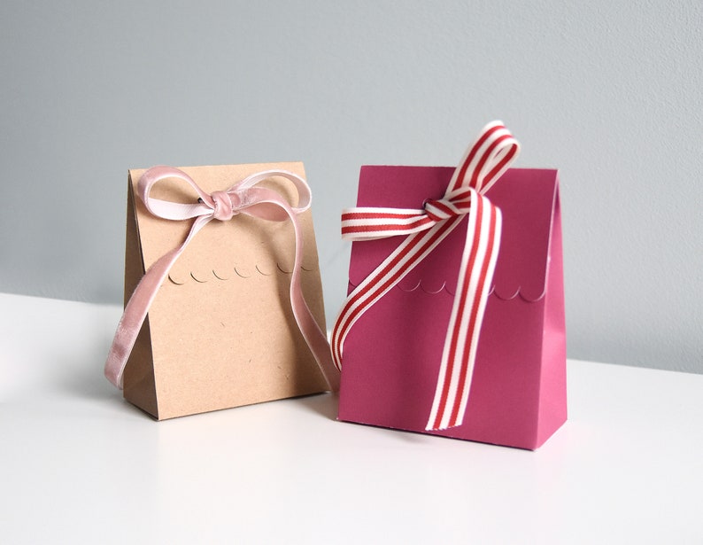 May include: Two small gift bags, one tan and one magenta, sit side-by-side. The tan bag has a pink velvet ribbon tied in a bow. The magenta bag has a red and white striped ribbon tied in a bow. Both bags have a scalloped edge.
