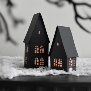 Peut inclure: Deux décorations de maison en papier noir avec des fenêtres découpées, posées sur un tissu blanc en forme de toile d'araignée. Les maisons ont des toits pointus et sont éclairées de l'intérieur, créant une lueur chaleureuse.