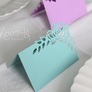 May include: A light blue place card with a delicate floral design cut out. The design features a large flower with leaves and smaller flowers. The card is folded in half and has a blank space for writing a name.