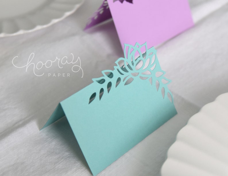May include: A light blue place card with a delicate floral design cut out. The design features a large flower with leaves and smaller flowers. The card is folded in half and has a blank space for writing a name.