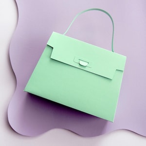 Pode incluir: Uma bolsa de papel verde-menta com uma alça curva e um fecho de aba retangular. A bolsa está sobre um fundo roxo suave e ondulado.