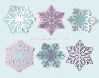 May include: Six paper snowflakes in various shades of blue, pink, and purple. The snowflakes are cut out of paper and have a layered effect. The snowflakes are arranged in a row on a light blue background. The text "HOORAYPAPER.COM" is printed below the snowflakes.