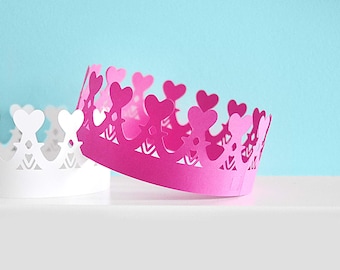 Op de afbeelding: Twee papieren kronen, een witte en een roze met een hartvormig ontwerp. De roze kroon is een verloop van roze en magenta.