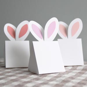 May include: Three white paper gift boxes with pink bunny ears. The boxes are on a brown and white checked tablecloth.