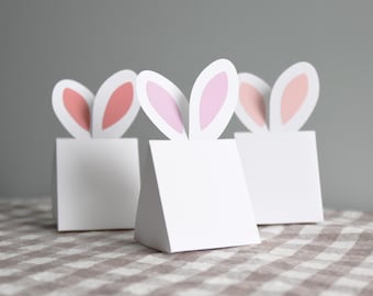 May include: Three white paper gift boxes with pink bunny ears. The boxes are on a brown and white checkered tablecloth.