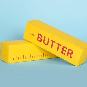 Könnte beinhalten: Zwei gelbe Butterboxen mit rotem Text und einem Lineal-Design. Eine Box ist abgewinkelt und zeigt das Wort "BUTTER" in Rot. Die andere Box befindet sich darunter, mit einem Lineal-Design und dem Text "4oz". Der Hintergrund ist hellblau.