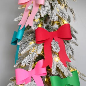 Può includere: Un albero di Natale decorato con fiocchi di carta colorati nelle tonalità del rosa, del rosso, del verde acqua e del verde. L'albero è innevato con neve artificiale e ha piccole luci bianche calde.