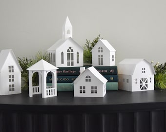 May include: A set of six white paper houses, including a church, a barn, a gazebo, and three houses. The houses are arranged on a black surface with books stacked behind them.