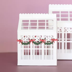 Puede incluir: Cajas blancas de papel con forma de invernadero y recortes de ventanas. La caja delantera tiene coronas verdes con lazos rojos. El fondo es de color rosa liso. Las cajas tienen bordes festoneados y probablemente son para fines decorativos.