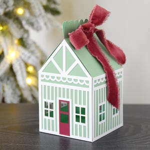 May include: A house-shaped gift box in green and white, with a red door and a matching red ribbon. The box has a scalloped roofline and vertical striped detailing. The box is designed to resemble a gingerbread house.