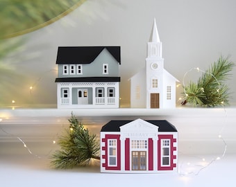 May include: Three paper houses: a gray house with a black roof, a white church with a steeple, and a red and white library. The miniature buildings are arranged on a white surface with greenery and string lights.