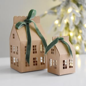 Puede incluir: Dos casas de papel marrón con ventanas recortadas y lazos de cinta de terciopelo verde. Las casas están sobre una superficie blanca frente a un fondo borroso de un árbol de Navidad con luces blancas.