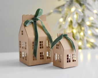 Puede incluir: Dos casas de papel marrón con ventanas recortadas y lazos de cinta de terciopelo verde. Las casas están sobre una superficie blanca frente a un fondo borroso de un árbol de Navidad con luces blancas.