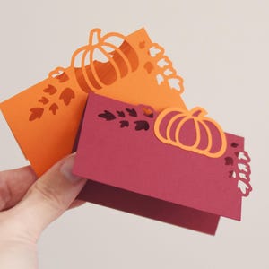 May include: Two fall-themed place cards. One is orange, the other is burgundy. Each card features a pumpkin and leaf cut-out design. The cards are held in a hand against a neutral background. Perfect for Thanksgiving or autumn events.