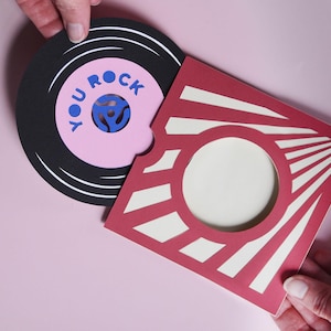 Könnte beinhalten: Eine Grußkarte aus Papier in Form einer Schallplattenhülle in Rot und Creme mit kreisförmigem Ausschnitt. Eine schwarze Schallplatte aus Papier mit rosa Mittellabel und dem blauen Text YOU ROCK wird teilweise aus der quadratischen Hülle gezogen.