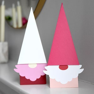 May include: Two paper gnome ornaments, one with a pink hat and the other with a red hat. Both gnomes have white beards and red noses.