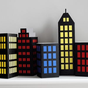 May include: A set of five paper cityscape buildings with black exteriors and colorful windows. The buildings are arranged in a row on a white surface. The windows are yellow, red, and blue.