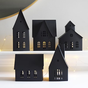Peut inclure: Cinq décorations de maison en papier noir avec différents styles de toits et des découpes de fenêtres. Les maisons sont disposées sur une surface blanche, avec une guirlande de petites lumières en arrière-plan. Les maisons sont éclairées de l'intérieur, créant une lueur chaleureuse.