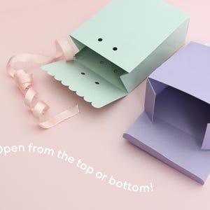 May include: Two pastel-colored gift boxes, one mint green and one lavender, are open on a pink surface. A pink ribbon is curled near the green box. The text "Open from the top or bottom!" is visible.