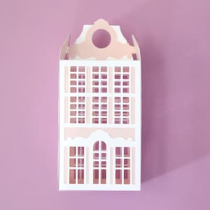 Pode incluir: Uma maquete de papel de uma casa em estilo holandês, com fachada branca e detalhes em rosa. O edifício tem inúmeras janelas e um topo decorativo com uma abertura circular. O fundo é rosa liso.