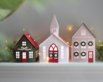 May include: Three decorative paper houses illuminated by thin string lights. The left house is dark green with a red roof, the center is a white church with a red door and steeple, and the right house is white with red stripes and green wreaths.