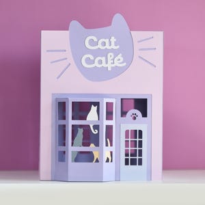 Op de afbeelding: Een lichtroze papieren model van een kattencafé. Het bord boven het café luidt "Cat Cafe" in witte letters. Het café heeft een lichtpaarse gevel met katten silhouetten in de ramen en deuren.