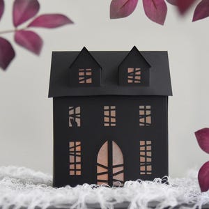 Op de afbeelding: Een zwart papieren huis met uitgesneden ramen en een deur, van binnenuit verlicht. Het huis heeft twee kleine dakkapellen op het dak. De scène wordt omlijst door paarse bladeren, wat een Halloween- of griezelthema creëert.