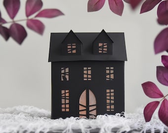 May include: A black paper house with cut-out windows and a door, lit from within. The house features two small dormers on the roof. The scene is framed by purple leaves, creating a Halloween or spooky theme.