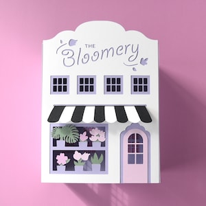 Pode incluir: Um recorte de papel de uma floricultura, "The Bloomery", com um toldo listrado em preto e branco e uma porta rosa. As janelas da loja exibem flores e plantas de papel. O fundo é rosa sólido.