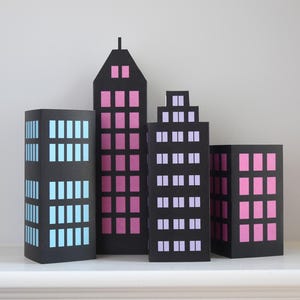 Könnte beinhalten: Eine Sammlung von vier schwarzen Papier-Stadtgebäuden mit farbenfrohen Fensterausschnitten. Die Gebäude variieren in Höhe und Form, mit rosa, blauen und lila Fenstern. Das höchste Gebäude hat ein spitzes Dach.