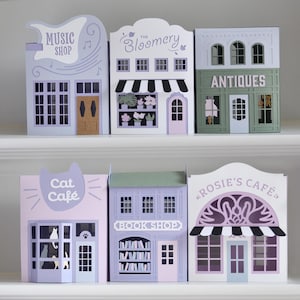 May include: Six decorative paper storefront models featuring Music Shop, The Bloomery, Antiques, Cat Café, Book Shop, and Rosie's Café. Each structure displays unique architectural details, signage, and colour schemes including lavender, white, and green.