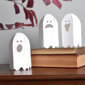 Op de afbeelding: Drie witte papieren spookdecoraties met verschillende gezichtsuitdrukkingen, geplaatst op oude boeken. Eén spook heeft een open mond, een ander een verdrietig gezicht en de derde een lachend gezicht. De spoken staan op een houten oppervlak.